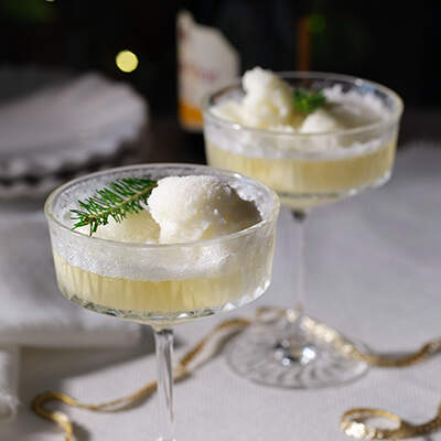 In einem flachen Coupeglas befindet sich eine helle Gelée mit einem Klecks Sorbet; ein grünes Zweigchen Thymian dient als Dekoration