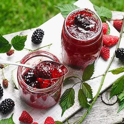 Auf einem weißen Brett steht ein offenes Glas mit tiefrotem Beerengelee, aus dem ein Löffel mit glänzendem Inhalt herausragt. Daneben steht ein weiteres Glas mit Deckel, frische Brombeeren und Himbeeren sowie grüne Blätter sind dekorativ verteilt.
