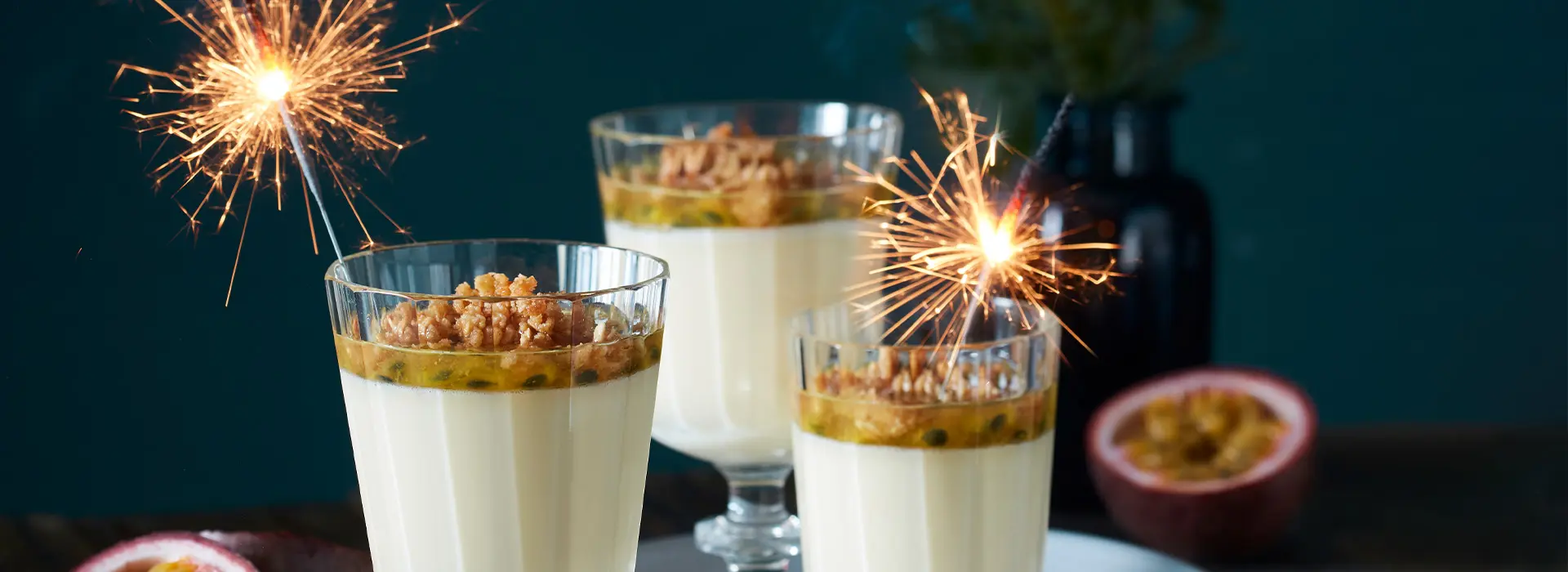 Drei hohe Gläser beinhalten eine helle Creme, darauf eine Schicht Marmelade und Nüsse. In zwei der drei Gläsern stecken funkelnde Wunderkerzen. 