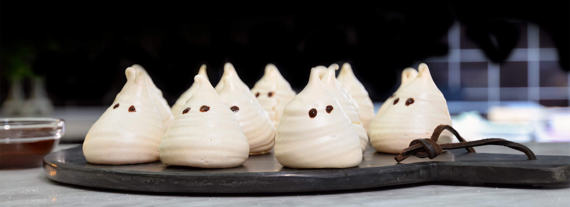 weiße Meringue-Geister in einer Reihe angeordnet. Jeder Geist hat eine charakteristische Spitzform mit zwei dunklen Schokoladenpunkten als Augen und einen dünnen, gewellten Schokoladenstrang als Mund.