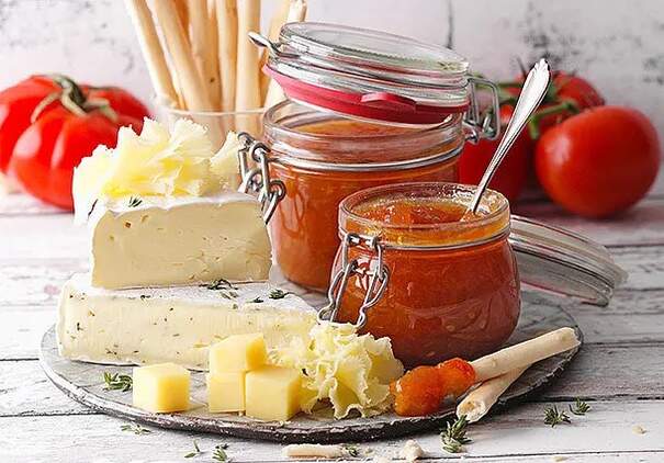 Auf einem Marmortablett ist ein appetitliches Arrangement aus mehreren Elementen zu sehen: Scheiben eines hellen, würzigen Käses, mehrere Gläser mit dunkelroter Marmelade oder Gelee, frische rote Tomaten, grüne Paprika, Streichhölzer zum Servieren und verstreute Rosmarin-Sprigs.