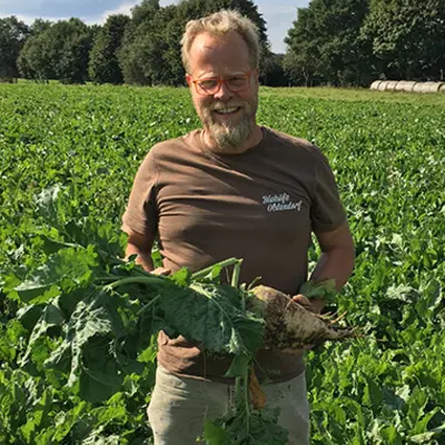 Ein Landwirt hält eine große Zuckerrübe in der Hand und steht vor einem großen grünen Feld voller Zuckerrüben