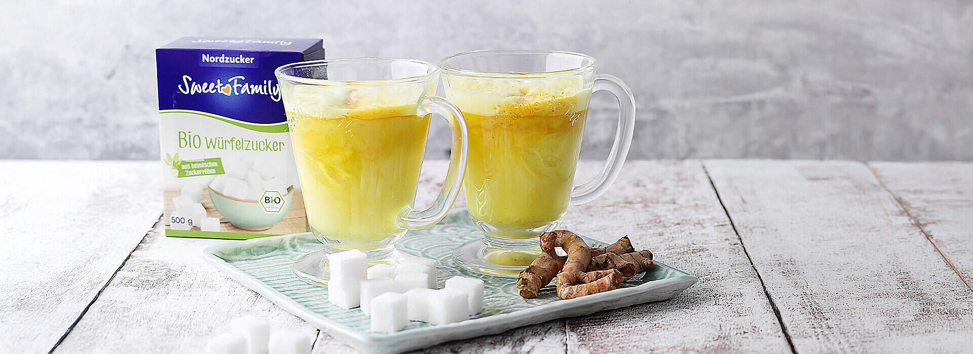 Zwei Glasbecher gefüllt mit leuchtend gelber Goldener Milch stehen auf einem hellen Holzbrett; davor liegen Zuckerwürfel und frische Kurkumawurzeln, im Hintergrund eine Packung Bio-Würfelzucker.