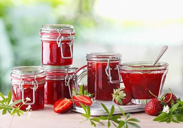 Mehrere unterschiedlich große Einweckgläser mit dunkelrotem Erdbeer‑Waldmeister‑Aufstrich stehen auf einer hellen Tischplatte. Ein Glas ist geöffnet, und ein Löffel ruht in der glänzenden Masse, während die anderen mit Metallbügeln verschlossen sind. Um die Gläser liegen frische Erdbeeren sowie grüne Waldmeistertriebe mit sternförmigen Blättern und zarten weißen Blüten.