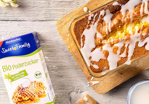 In einer Backform befindet sich ein hellbrauner Kuchen mit weißer Zuckerglasur; daneben steht eine Packung Bio-Puderzucker, die den Einsatz pflanzlicher Zutaten betont.