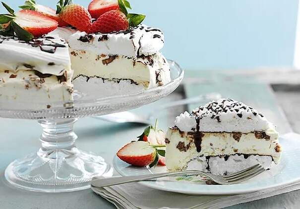 Eine üppige Meringue‑Eistorte mit Sahne, Schokostücken und Erdbeerhälften steht auf einer Kuchenplatte, während ein großes Stück daneben auf einem Teller liegt.