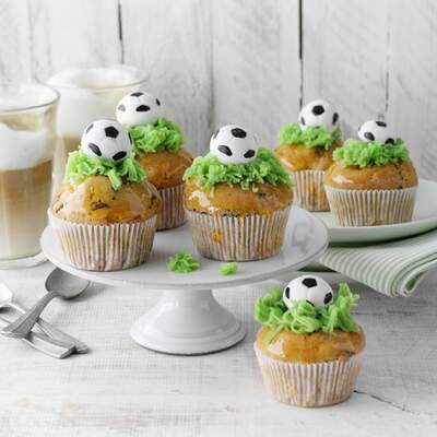 Auf einem Kuchenständer stehen Muffins aus hellem Teig mit grünem Frosting dekoriert. Obendrauf ist ein Fußball aus Zuckerguss