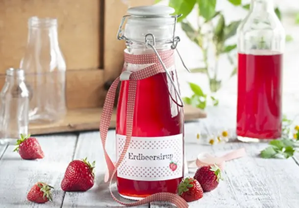 Glasflasche mit tiefrotem Erdbeersirup, beschriftetes Etikett und frische Erdbeeren dekorativ daneben