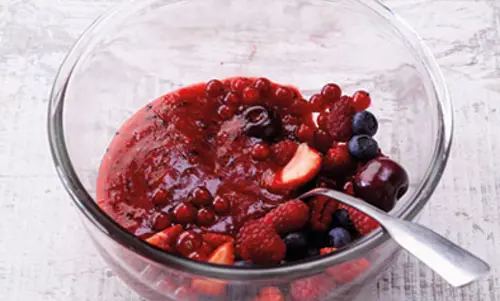 Eine große Glasschale beinhaltet Fruchte, eine Pfütze Fruchtsaft und einen großen Löffel. 