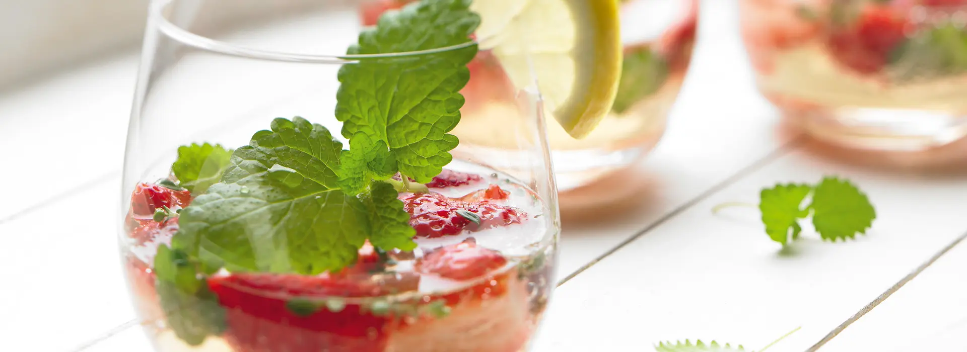 Ein heller Cocktail in einem bauchigen tiefen Glas, mit Himbeeren und Minze verziert. 