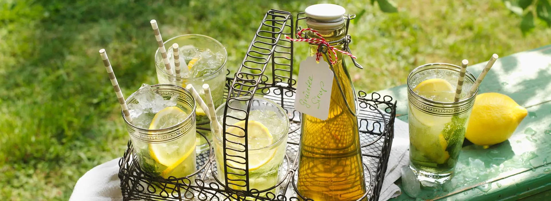 Auf einem verspielten Tablet mit Henkel stehen drei Gläser mit Limonade und eine Glasflasche Sirup.