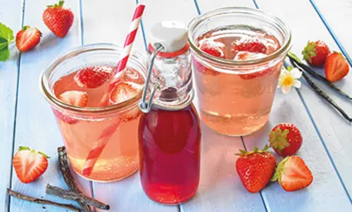 Schritt 5 In zwei breiten Gläser, die ebenso gut Einmachgläser sein könntent, ist eine hell-rosane Limonade enthalten, in der Erdbeeren und ein rot gestreifter Strohhalm schwimmen. Davor steht eine kleine Glasflasche mit Sirup drin.