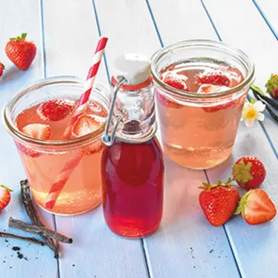 selbstgemachter Erdbeersirup steht in zwei Gläsern und einer Flasche auf einem blauen Holztisch. Daneben liegen frische Erdbeeren