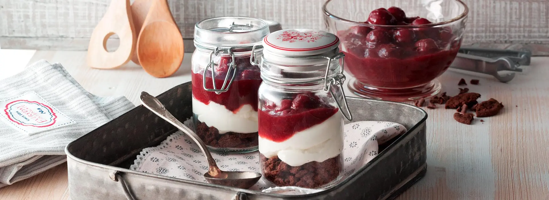 Auf einem Tablet stehen zwei hohe Einmachgläser, die Sckokokekse, Mascarpone und Kirschkompott beinhalten.
