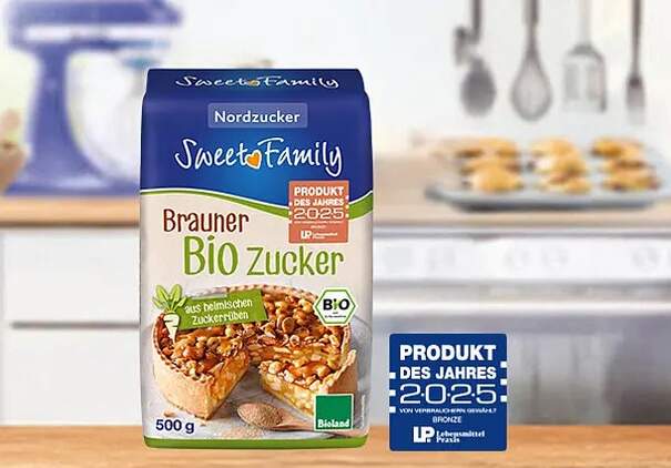 Auf dem Bild steht ein Päckchen Zucker der Marke Sweet Family (Nordzucker). Es handelt sich dabei um braunen Bio-Zucker. Der Zucker steht auf einer Arbeitsplatte in der Küche. Auf dem Zuckerpäckchen ist noch ein Kuchen abgebildet. Daneben sieht man das Siegel "Produkt des Jahres 2025". 