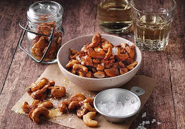 Auf einem rustikalen Holztisch steht eine gesprenkelte Keramikschale, gefüllt mit glänzenden, karamellisierten Cashewkernen.
