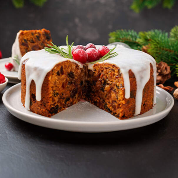 Auf einem dunklen Untergrund steht ein runder, dunkler Früchtekuchen auf einer weißen Platte. Ein Stück wurde bereits herausgeschnitten, wodurch die saftige, mit Früchten und Nüssen gefüllte Textur sichtbar wird. Der Kuchen ist mit einer dicken, weißen Glasur überzogen, die an den Seiten herunterläuft.