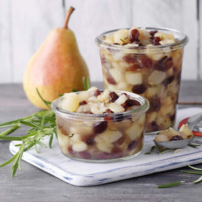 Zwei Gläser mit Birnenkompott und Preiselbeeren, umgeben von frischen Birnen und Cranberries.