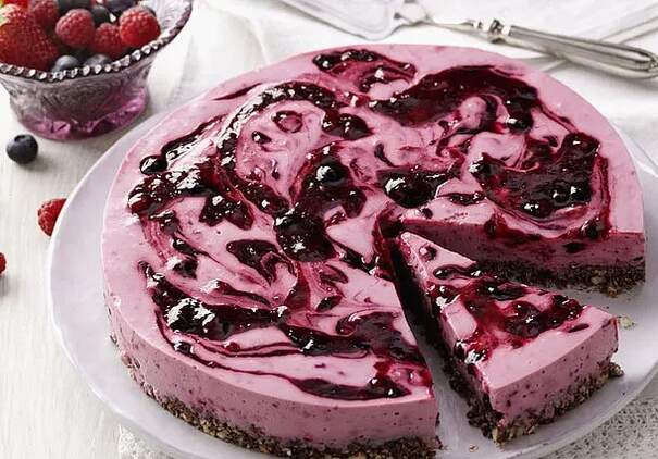 Eine cremige Torte mit weißem Belag und dunkler Rote‑Grütze‑Spirale steht auf einer flachen Platte; ein Stück ist bereits herausgeschnitten.