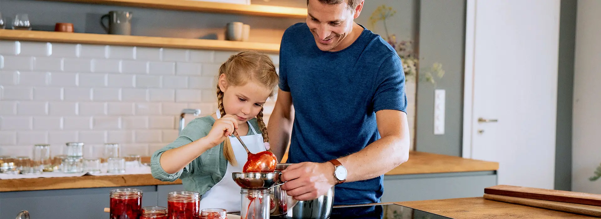 Eine Vater‑Tochter‑Szene in der Küche: beide kochen Marmelade, mehrere Gläser stehen bereit.