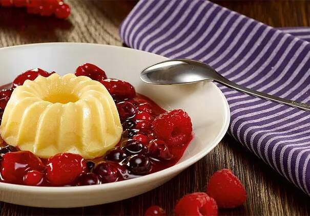 In einem tiefen weißen Teller liegt eine hellgelbe Vanillepuddingform in Gugelhupfform, umgeben von einer glänzenden, dunkelroten Beerenkompott. Einzelne Himbeeren und Johannisbeeren schwimmen in der Sauce und glänzen im Licht.