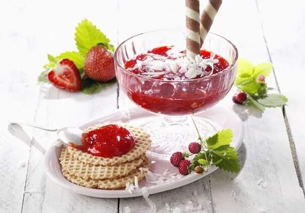 Ein Glas roter Fruchtaufstrich steht in einem Korb, daneben Waffeln, Erdbeeren und Blaubeeren.