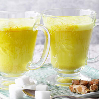 Ein Glas goldgelbe Kurkuma‑Milch steht mit Milchschaum und Zimt bestäubt neben einem Stück Ingwer.