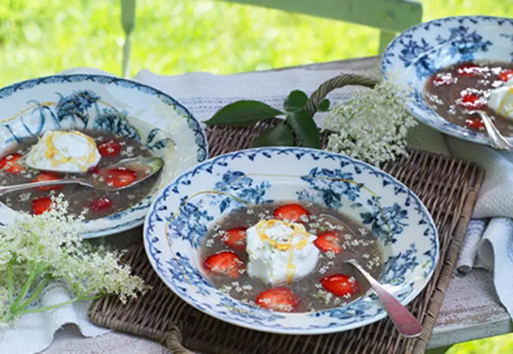 Zwei blaue Schalen mit klarer Holunderblüten‑Kaltschale, darin schwimmen Erdbeeren und luftige Schneeeier, garniert mit Blüten