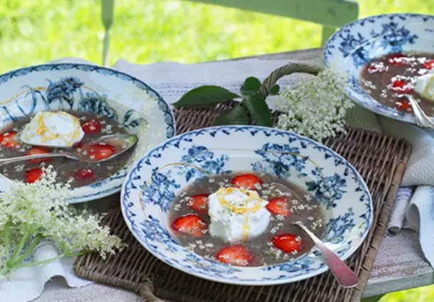 Zwei blaue Schalen mit klarer Holunderblüten‑Kaltschale, darin schwimmen Erdbeeren und luftige Schneeeier, garniert mit Blüten