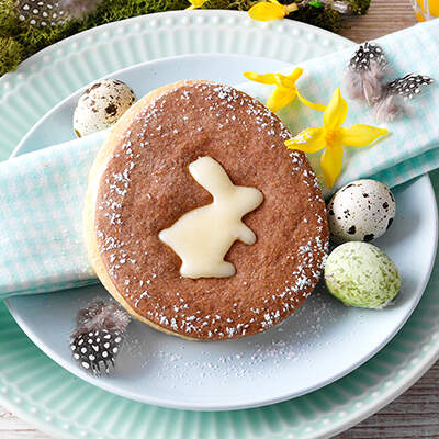 Auf einem türkisfarbenen Teller steht ein heller Kuchen mit Zuckerglasur; darauf ist ein weißes Häschenmotiv aus Puderzucker gestreut, umgeben von kleinen Wachteleiern und gelben Narzissen
