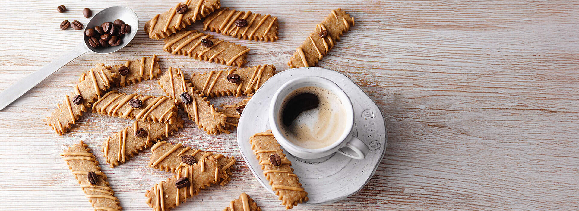 Eine Tasse Kaffee mit dichter Crema steht auf einer Untertasse; umgeben wird sie von rechteckigen Keksen mit Zuckerguss sowie verstreuten Kaffeebohnen auf einer hellen Holzoberfläche.
