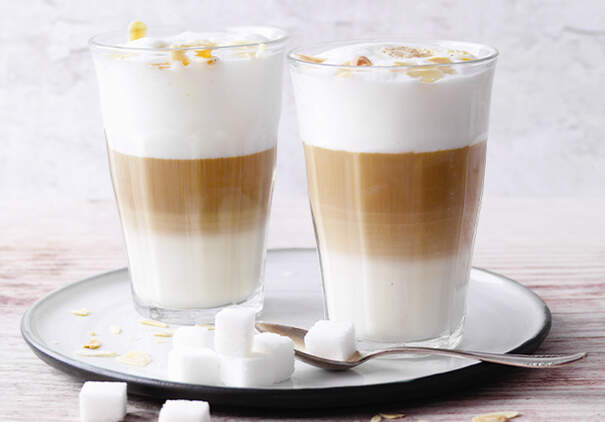 Zwei hohe Gläser mit Latte macchiato zeigen klar abgesetzte Schichten aus Espresso, Milch und Schaum; daneben stehen Zuckerstücke auf einem Teller.