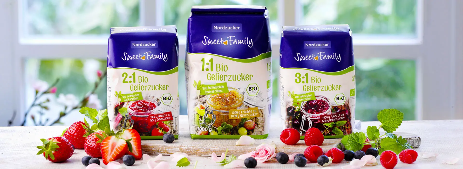 Drei verschiedene Bio-Zucker Produkte von Sweetfamily sind nebeneinander aufgestellt. Umringt von frischen Beeren.