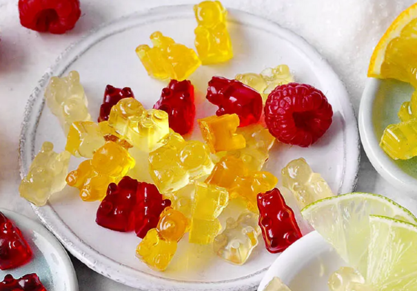 Auf einem kleinen weißen Teller liegen zahlreiche gelbe und rote Gummibärchen dicht beieinander, dazwischen liegt eine frische Himbeere. Um den Teller herum sind Scheiben von Limette und Zitrone sowie weitere Bärchen dekorativ verteilt.