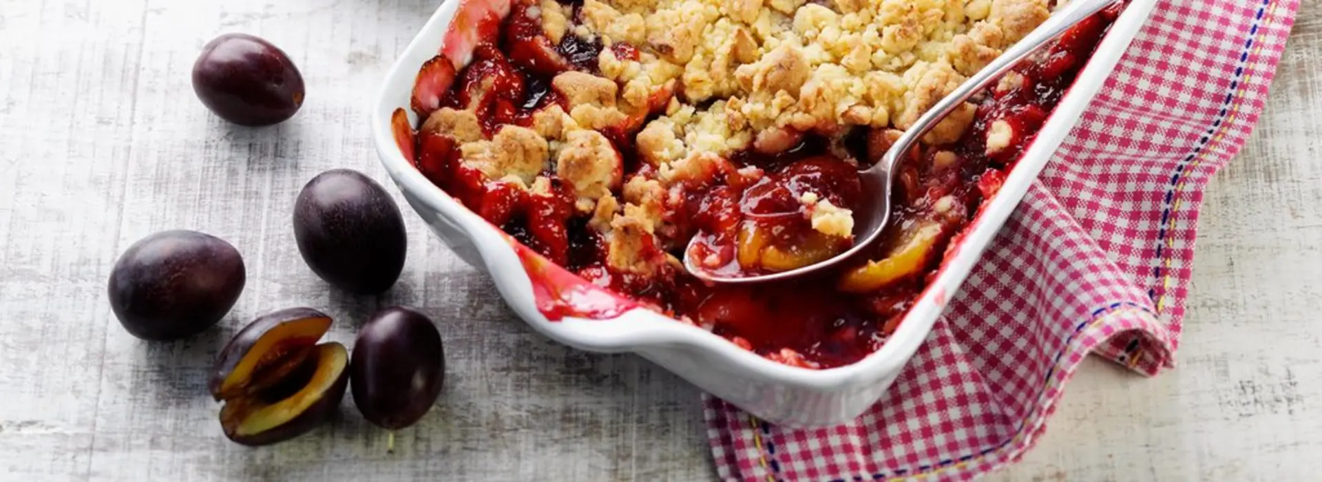 Eine weiße Auflaufform mit Pflaumen‑Crumble ist mit Streuseln bedeckt; daneben liegen frische Pflaumen.