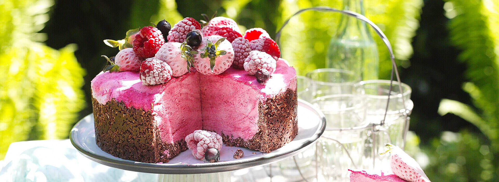 Auf einer weißen Tortenplatte thront eine mehrschichtige Beeren‑Eistorte in zartem Rosa, bedeckt mit gefrorenen Himbeeren, Brombeeren und Erdbeerscheiben. Die Oberfläche glitzert leicht, da die Früchte mit Eiskristallen überzogen sind, und grüne Blätter setzen frische Akzente.