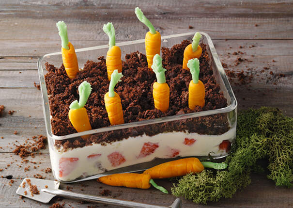 In einer rechteckigen Glasschale sind mehrere Lagen einer Dessertcreme geschichtet; darauf liegen Marzipankarotten, die in dunkle Keksbrösel gesteckt sind und wie ein kleiner Gemüsegarten wirken.