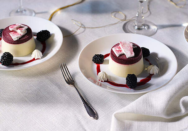 Zwei kleine runde Desserts bestehen aus einem hellen Panna-cotta‑Sockel und einem dunklen Fruchtspiegel; sie sind mit Beeren und Meringuetupfen dekoriert