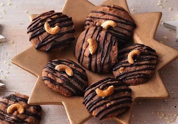 Auf einem sternförmigen Teller liegen runde braune Cookies. Sie sind mit Schokoladenstreifen überzogen und je mit einer Cashewnuss in der Mitte verziert. 