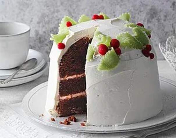 Auf einer weißen Tortenplatte mit filigranem Rand steht eine hohe, runde Schokoladentorte, die vollständig mit einer glatten, weißen Creme überzogen ist. Ein großzügiges Stück wurde herausgeschnitten und enthüllt zwei dunkle, saftige Kuchenböden, die durch eine feine, rötliche Cremeschicht getrennt sind. Die Torte ist mit hellgrünen, blattförmigen Verzierungen und kleinen, leuchtend roten Beeren dekoriert.