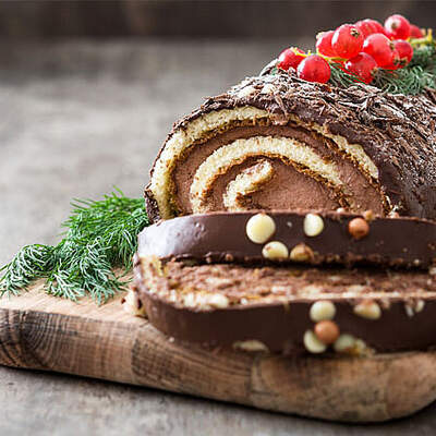 Eine schokoladige Biskuitrolle mit spiralförmiger Cremefüllung liegt auf einem Holzbrett; sie ist mit Glanzglasur überzogen und mit kleinen Goldperlen, Cranberries und Tannenzweigen dekoriert.
