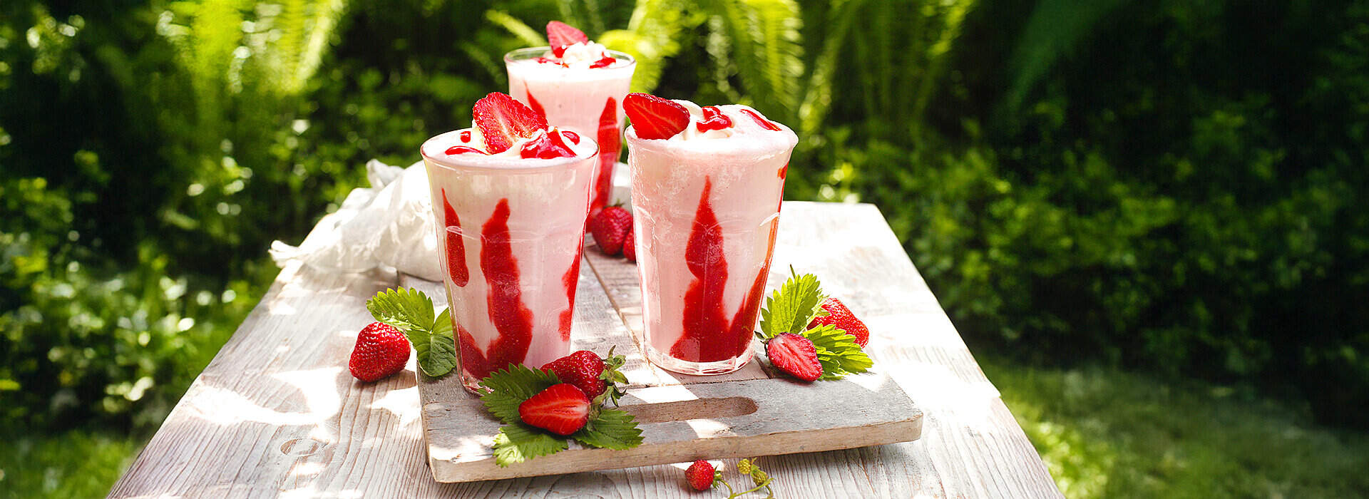 Auf einem rustikalen Brett stehen drei Glasbecher mit rosa gefrorenem Erdbeer‑Frappuccino, jeder mit einer dekorativen Sahnehaube.