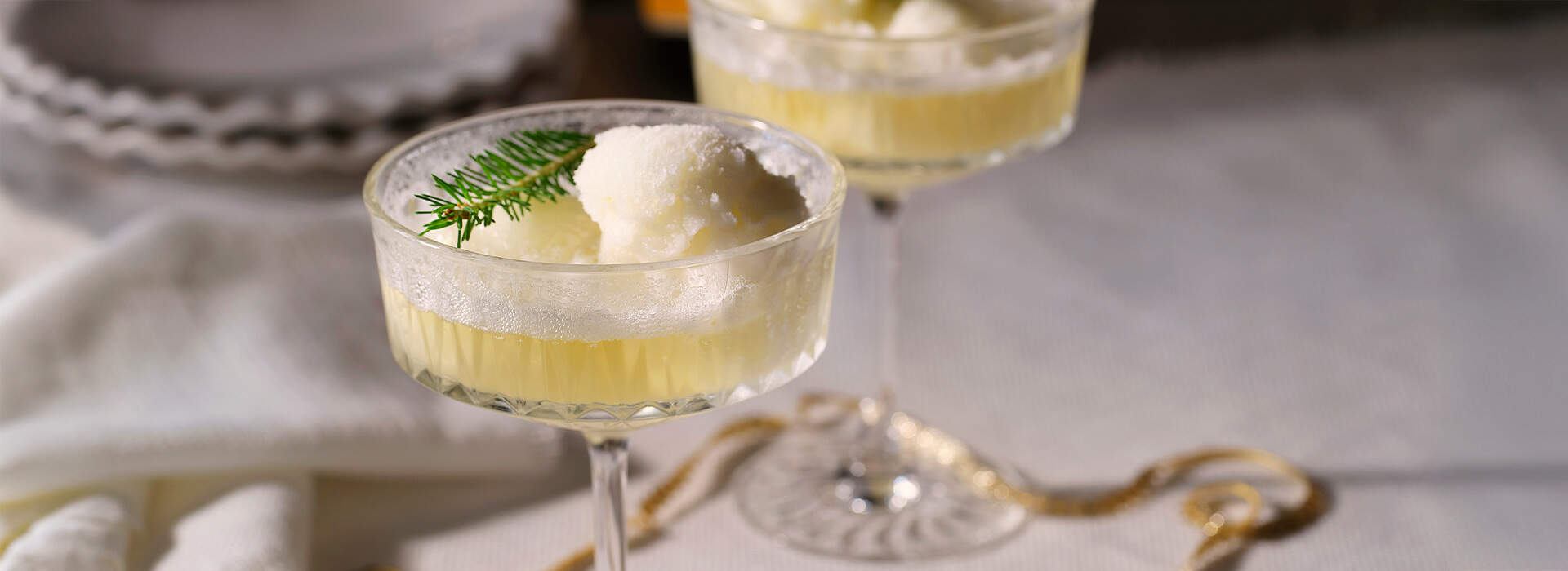 Auf dem Bild sieht man ein hohes, rundes Cocktailglas, das zur Hälfte mit einem gelben Cocktail gefüllt ist. Im Glas findet sich zudem eine Kugel Eis und ein kleiner Tannenzweig liegt dekorativ darüber. 