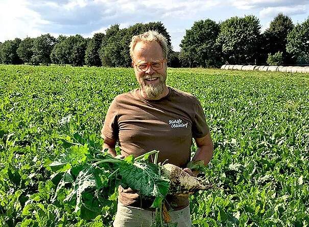 Ein älterer Mann mit Brille und grauem Bart steht in einem Zuckerrübenfeld und hält stolz eine Zuckerrübe mit Blättern in den Händen. Er trägt ein braunes T-Shirt mit einem Logo und lächelt in die Kamera. Im Hintergrund erstreckt sich das grüne Feld unter einem blauen Himmel mit weißen Wolken.