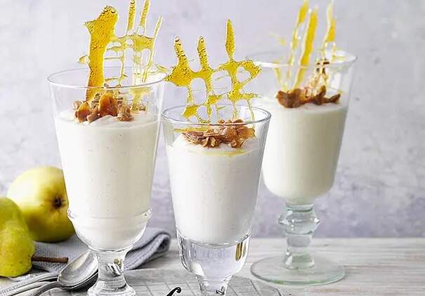 Drei hohe Dessertgläser sind bis zum Rand mit einer luftigen, weißen Mousse gefüllt und stehen auf einem hellen Holztisch. Auf jedem Glas liegen karamellisierte Nüsse, aus denen goldene Zuckerspitzen wie filigrane Skulpturen herausragen.