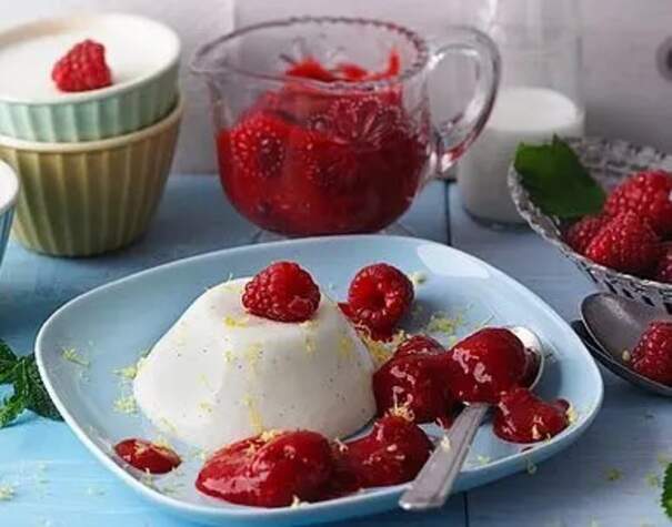 Auf einem hellblauen Teller liegt ein weißer Panna‑Cotta‑Halbkegel, der mit rotem Himbeermark und frischen Himbeeren übergossen ist.