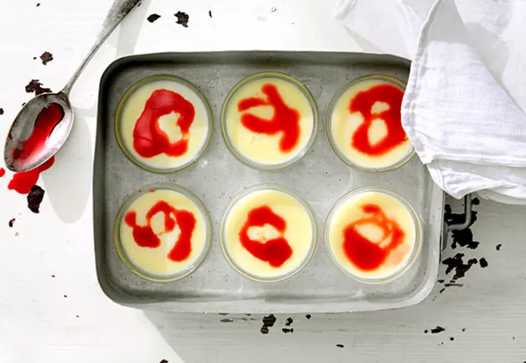 Weiße Panna Cotta steht in Gläsern und wird von leuchtend rotem Erdbeersirup überzogen, dazu einige frische Erdbeeren