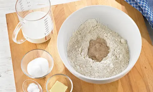 Schritt 1 Neben einer Schüssel mit Mehl steht ein Messbecher Milch, eine Schale Zucker, Butter und Backstärke
