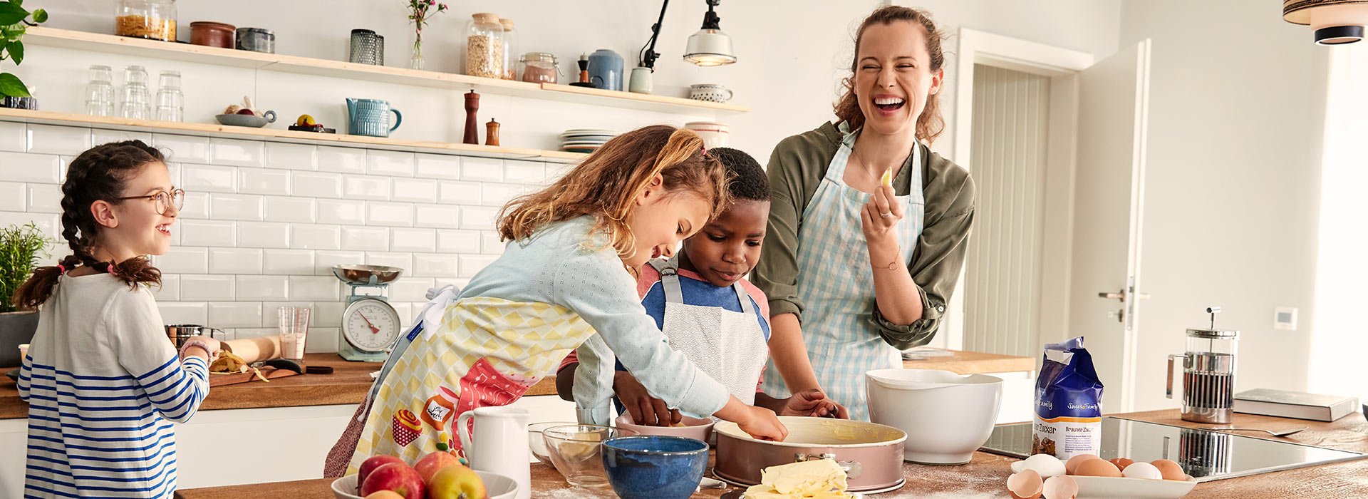 Eine Frau backt mit drei Kinden einen Kuchen. Sie freuen sich. Sie verwenden Produkte von SweetFamily