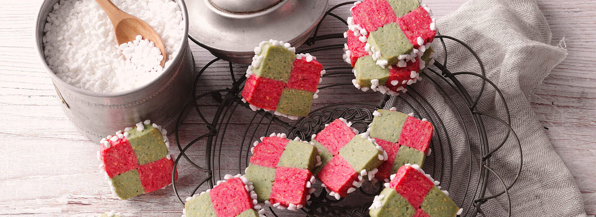 Bunte quadratische Plätzchen liegen eng aneinander auf einer grauen Platte. Die Oberflächen zeigen rote und grüne Muster und sind mit Zucker bestreut. Rechts daneben steht ein Glasröhrchen mit Zucker, dahinter liegt ein gefaltetes Leinentuch.